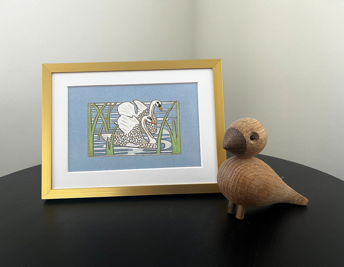 Kunstkaart met zwanen ingelijst in een gouden fotolijst met een houten beeldje van een vogel ernaast. 